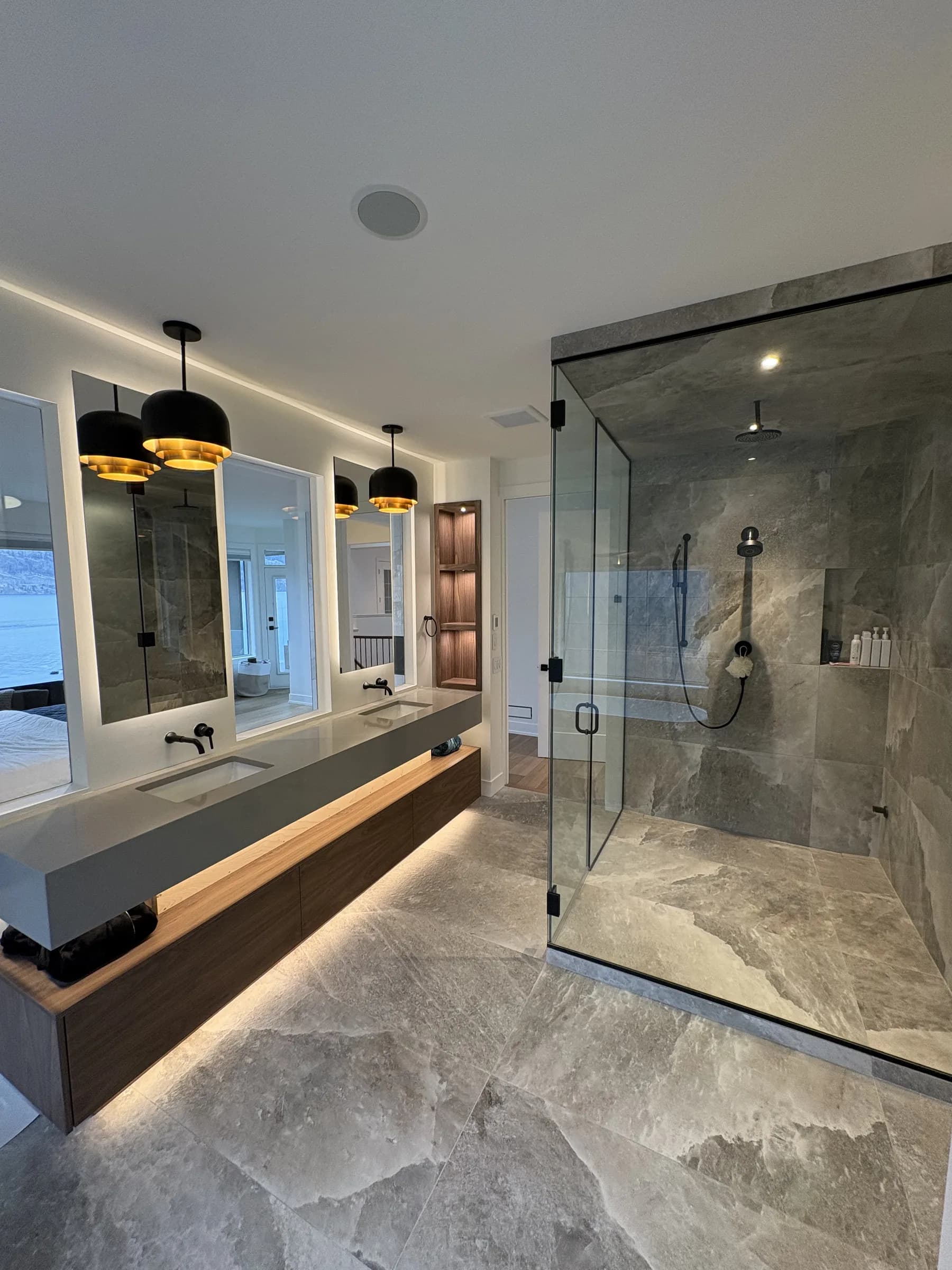 Stone-tiled ensuite with a glass steam shower, underlit floating vanity and black-and-brass pendants.