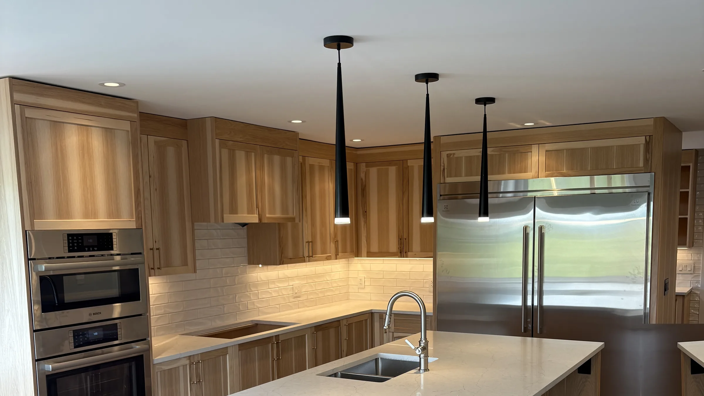 Bright custom kitchen in light oak with black cone pendants, under-cabinet lighting and panel-ready appliances.